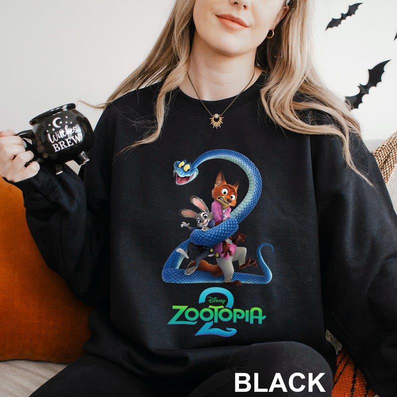 May include: Black sweatshirt featuring a colorful graphic from the Disney movie Zootopia 2. The design includes the characters Nick Wilde, Judy Hopps, and a blue snake. The word "Zootopia 2" is in a teal font, and the word "BLACK" is at the bottom.