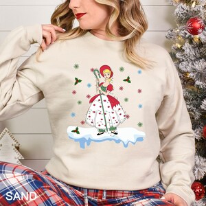 Puede incluir: Sudadera color arena con una ilustraci&oacute;n de dibujos animados de una ni&ntilde;a con un vestido rojo y blanco, sosteniendo un bast&oacute;n verde. El dise&ntilde;o incluye copos de nieve y acentos de acebo, evocando un tema de vacaciones de invierno.