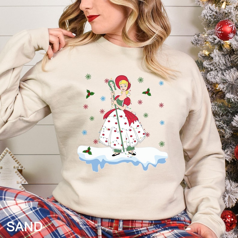 Puede incluir: Sudadera color arena con una ilustraci&oacute;n de dibujos animados de una ni&ntilde;a con un vestido rojo y blanco, sosteniendo un bast&oacute;n verde. El dise&ntilde;o incluye copos de nieve y acentos de acebo, evocando un tema de vacaciones de invierno.