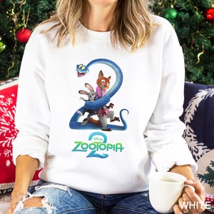 May include: White sweatshirt featuring a colorful graphic of characters from the animated movie Zootopia 2. The design includes a blue snake wrapped around a fox and a bunny, with the movie title in green and blue.