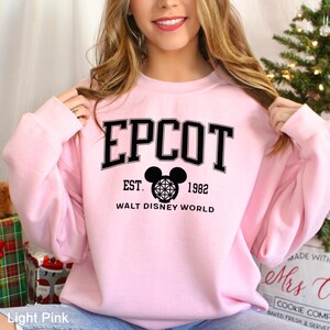 K&ouml;nnte beinhalten: Hellrosa Sweatshirt mit dem Wort "EPCOT" in schwarzen Blockbuchstaben. Darunter steht der Text "EST. 1982 WALT DISNEY WORLD" in Schwarz. Ein Mickey Mouse Kopf-Logo ist unter dem Wort "EPCOT" zentriert.