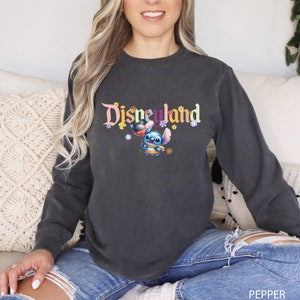 Puede incluir: Una sudadera gris oscuro de manga larga con el texto "Disneyland" en letras de colores. Un personaje azul Stitch sostiene un globo de Mickey Mouse delante del texto.