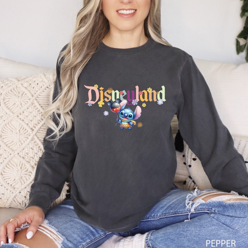 Puede incluir: Una sudadera gris oscuro de manga larga con el texto "Disneyland" en letras de colores. Un personaje azul Stitch sostiene un globo de Mickey Mouse delante del texto.