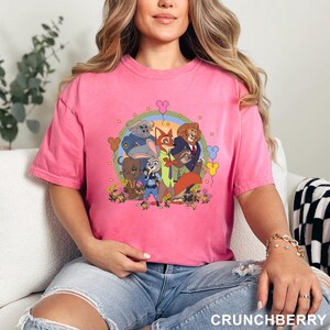 May include: A pink t-shirt featuring a colorful graphic of characters from the animated movie "Zootopia". The design includes Judy Hopps, Nick Wilde, and other characters, surrounded by flowers and a rainbow. The text "CRUNCHBERRY" is at the bottom.