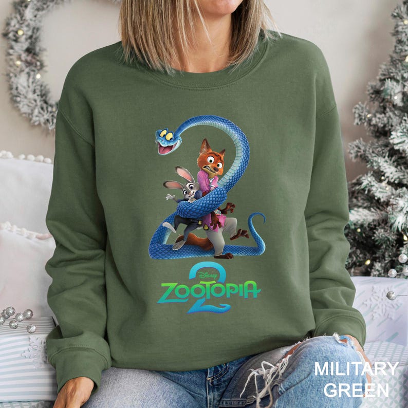 May include: Military green sweatshirt featuring a graphic of characters from the animated movie Zootopia 2. The design includes a blue snake, a fox, and a rabbit. The Zootopia 2 logo is displayed in teal and white.