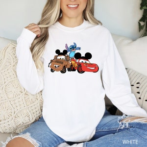 May include: White long-sleeved sweatshirt with a graphic print of Stitch, Mickey Mouse, and Mater from Disney's Cars. The characters are in a playful pose, with Stitch on top of Mater and Mickey Mouse driving a red race car.