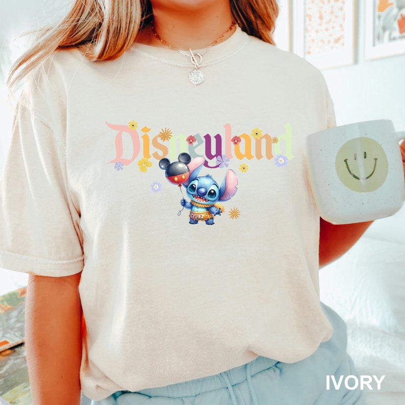 Puede incluir: Una camiseta blanca con el texto "Disneyland" en una fuente colorida. Un personaje azul Stitch sostiene un globo Mickey Mouse rojo y negro. La camiseta est&aacute; puesta en una modelo que lleva pantalones azules.