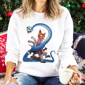 May include: White sweatshirt featuring a colorful graphic of characters from a popular animated movie. The design includes a blue snake wrapped around a fox and a rabbit. The sweatshirt is a comfortable, casual style.