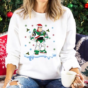 Puede incluir: Sudadera blanca con un personaje de dibujos animados de Buzz Lightyear con gorro de Pap&aacute; Noel y luces navide&ntilde;as. El dise&ntilde;o incluye copos de nieve y acebo. Un gr&aacute;fico festivo con tem&aacute;tica navide&ntilde;a.