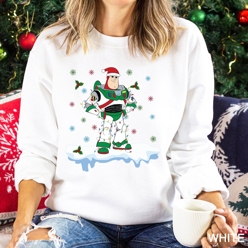 Puede incluir: Sudadera blanca con un personaje de dibujos animados de Buzz Lightyear con gorro de Pap&aacute; Noel y luces navide&ntilde;as. El dise&ntilde;o incluye copos de nieve y acebo. Un gr&aacute;fico festivo con tem&aacute;tica navide&ntilde;a.