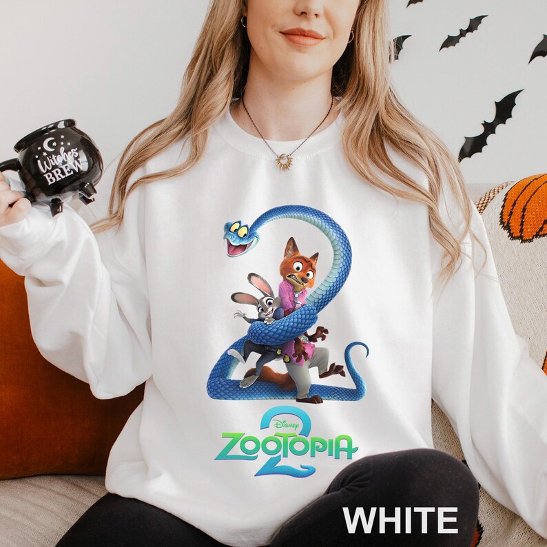 May include: White sweatshirt featuring a colorful graphic of characters from the movie Zootopia 2, including a fox, rabbit, and a blue snake. The text "Zootopia 2" is displayed in green and blue. The word "WHITE" is printed at the bottom.