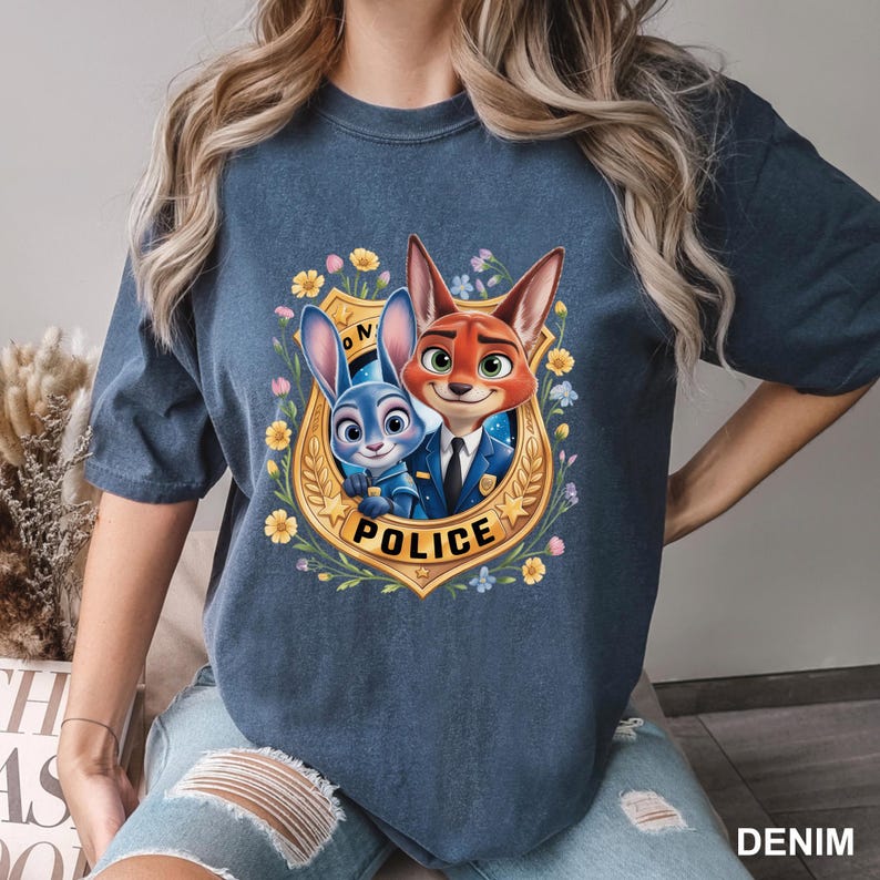 K&ouml;nnte beinhalten: Ein denimblaues T-Shirt mit einer farbenfrohen Grafik der Zootopia-Charaktere Judy Hopps und Nick Wilde in einem Polizeiausweis. Die Auszeichnung ist von gelben Blumen und gr&uuml;nen Bl&auml;ttern umgeben. Das Wort "POLICE" ist auf dem Ausweis aufgedruckt.
