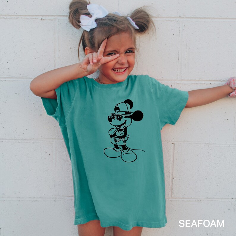 Może przedstawiać: Turkusowa koszulka z czarno-białym nadrukiem przedstawiającym Myszki Miki w okularach przeciwsłonecznych i czapce. W prawym dolnym rogu widoczny jest napis "SEAFOM".