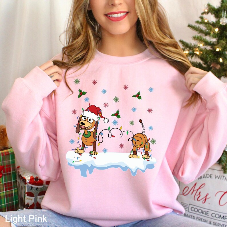 Puede incluir: Sudadera rosa claro con un perro de dibujos animados con gorro de Pap&aacute; Noel y luces navide&ntilde;as. El perro est&aacute; sobre una superficie cubierta de nieve, con copos de nieve y ramitas de acebo. El texto "Light Pink" est&aacute; en la parte inferior.