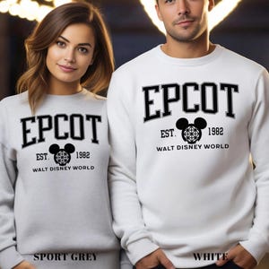 K&ouml;nnte beinhalten: Zwei Sweatshirts, eines in Sportgrau und eines in Wei&szlig;, jeweils mit dem Wort "EPCOT" in fetten schwarzen Buchstaben. Darunter eine Mickey-Mouse-Kopf-Grafik und der Text "EST. 1982 WALT DISNEY WORLD". Das Sportgrau-Sweatshirt hat unten den Text "SPORT GREY".