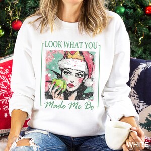 May include: White sweatshirt with a graphic of a woman wearing a Santa hat and holding a green apple. The text "LOOK WHAT YOU" and "Made Me Do" are printed in green. The design is framed in a green rectangle.
