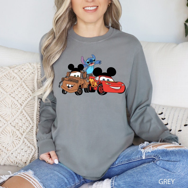 May include: A gray long-sleeve shirt with a graphic of Stitch, Mater, and Lightning McQueen from Disney's Cars and Lilo & Stitch. The shirt has a crew neck and long sleeves.