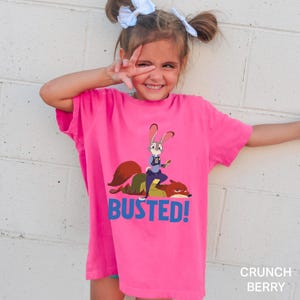 May include: A bright pink t-shirt featuring a cartoon image of a rabbit and fox with the word "BUSTED!" in blue. The shirt is a solid color and has short sleeves. The text "CRUNCH BERRY" is in the bottom right corner.