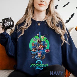 May include: Navy blue sweatshirt featuring a colorful graphic from the Disney movie Zootopia 2. The design includes various animal characters and the movie's logo. The word "NAVY" is printed at the bottom. A black mug with "Witches Brew" is held in the image.