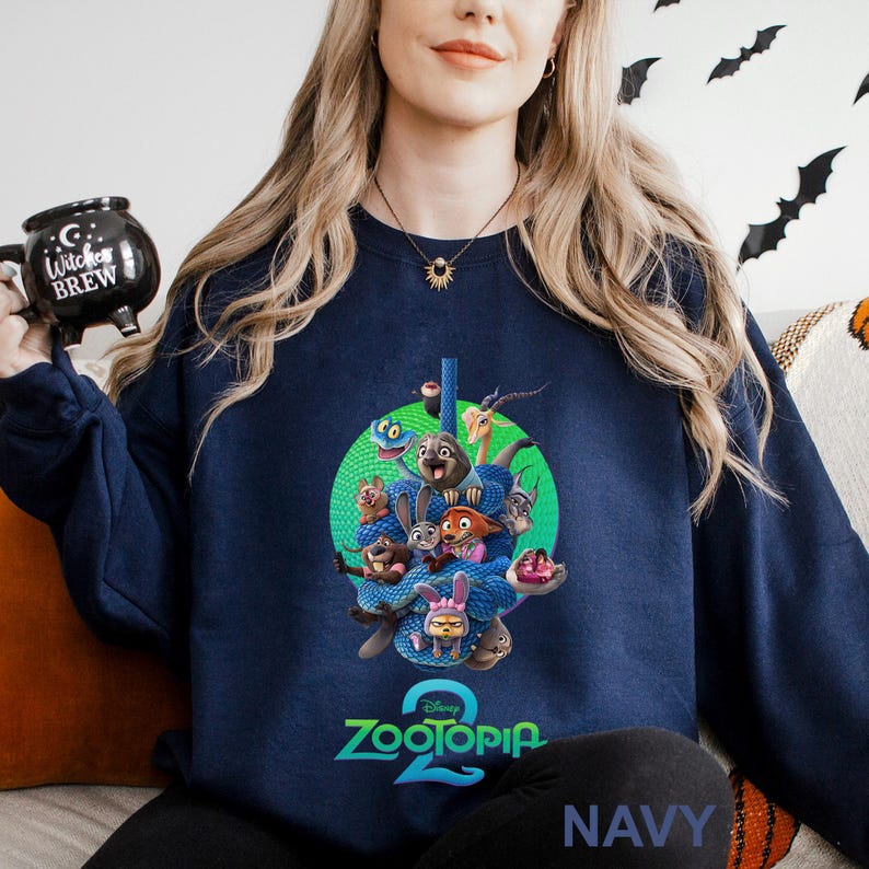 May include: Navy blue sweatshirt featuring a colorful graphic from the Disney movie Zootopia 2. The design includes various animal characters and the movie's logo. The word "NAVY" is printed at the bottom. A black mug with "Witches Brew" is held in the image.