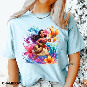 Puede incluir: Camiseta azul claro con una ilustraci&oacute;n vibrante de un personaje de dibujos animados tocando el ukelele. El dise&ntilde;o incluye flores coloridas, salpicaduras de agua y la palabra "CHAMBRAY". El personaje lleva una flor en el pelo y una falda tradicional.