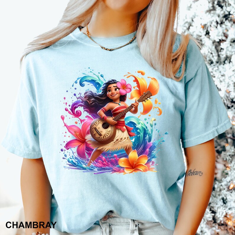 Puede incluir: Camiseta azul claro con una ilustraci&oacute;n vibrante de un personaje de dibujos animados tocando el ukelele. El dise&ntilde;o incluye flores coloridas, salpicaduras de agua y la palabra "CHAMBRAY". El personaje lleva una flor en el pelo y una falda tradicional.
