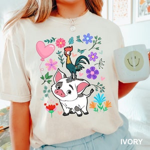 K&ouml;nnte beinhalten: Ein wei&szlig;es T-Shirt mit einem Cartoon-Bild eines Schweins und eines Hahns. Das Schwein ist wei&szlig; mit schwarzen Flecken und der Hahn ist rot, gelb und schwarz. Das Bild ist von bunten Blumen und Bl&auml;ttern umgeben. Der Text "IVORY" befindet sich in der rechten unteren Ecke des Bildes.