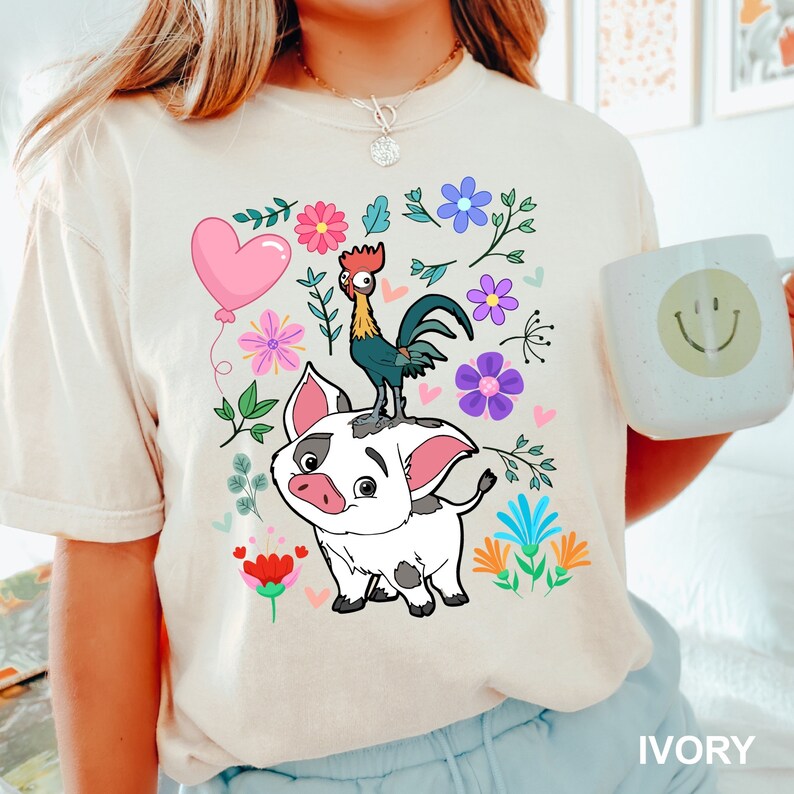 K&ouml;nnte beinhalten: Ein wei&szlig;es T-Shirt mit einem Cartoon-Bild eines Schweins und eines Hahns. Das Schwein ist wei&szlig; mit schwarzen Flecken und der Hahn ist rot, gelb und schwarz. Das Bild ist von bunten Blumen und Bl&auml;ttern umgeben. Der Text "IVORY" befindet sich in der rechten unteren Ecke des Bildes.