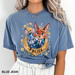 K&ouml;nnte beinhalten: Blaues Jeans-T-Shirt mit einem Cartoon-Bild eines Fuchses und eines Kaninchens in Polizeiuniformen. Das Bild befindet sich in einem goldenen Abzeichen mit dem Wort "POLICE" und ist von Blumen umgeben. Der Text "BLUE JEAN" befindet sich unten.