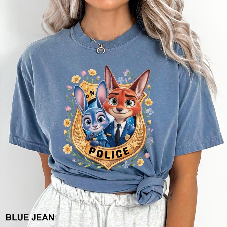 K&ouml;nnte beinhalten: Blaues Jeans-T-Shirt mit einem Cartoon-Bild eines Fuchses und eines Kaninchens in Polizeiuniformen. Das Bild befindet sich in einem goldenen Abzeichen mit dem Wort "POLICE" und ist von Blumen umgeben. Der Text "BLUE JEAN" befindet sich unten.