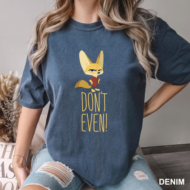 May include: A denim blue t-shirt featuring a cartoon fox with an angry expression, wearing a red vest. The text "DON'T EVEN!" is printed in yellow below the fox. The shirt is a casual wear item.