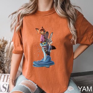 May include: Orange t-shirt featuring a graphic of characters from the animated movie Zootopia. The design includes Nick Wilde, Judy Hopps, and a blue snake. The shirt has short sleeves and a crew neck.