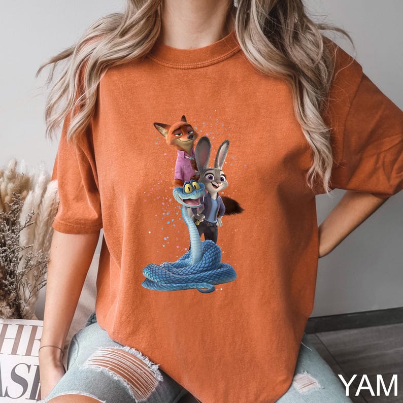 May include: Orange t-shirt featuring a graphic of characters from the animated movie Zootopia. The design includes Nick Wilde, Judy Hopps, and a blue snake. The shirt has short sleeves and a crew neck.