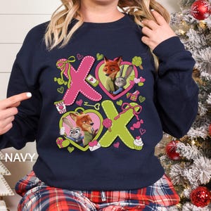 May include: Navy blue sweatshirt featuring a graphic design with the characters Nick Wilde and Judy Hopps from Zootopia. The design includes pink and green hearts, the letters "X" and "O", and other Valentine's Day themed elements.