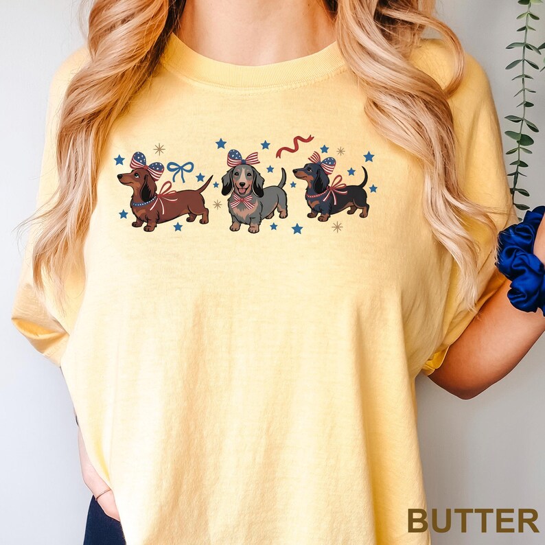 May include: Yellow t-shirt featuring three dachshunds with patriotic bows and star accents. The dogs are brown, gray, and black, with red, white, and blue bows. The word "BUTTER" is printed at the bottom of the shirt.