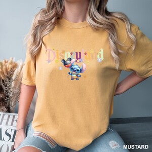 Puede incluir: Una camiseta amarilla mostaza con un gr&aacute;fico de "Disneyland" colorido que presenta a Stitch sosteniendo un globo de Mickey Mouse.