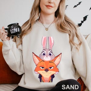 May include: Sand-colored sweatshirt featuring a graphic of a fox and a rabbit from a popular animated movie. The fox is orange with a smug expression, and the rabbit is gray with pink ears. The sweatshirt is a neutral color, suitable for casual wear.
