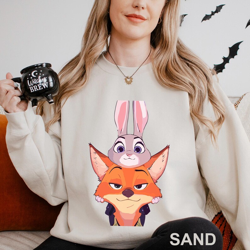 May include: Sand-colored sweatshirt featuring a graphic of a fox and a rabbit from a popular animated movie. The fox is orange with a smug expression, and the rabbit is gray with pink ears. The sweatshirt is a neutral color, suitable for casual wear.