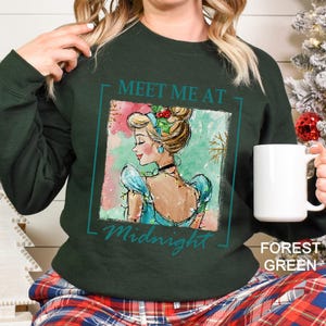May include: Forest green sweatshirt with a Cinderella graphic. The graphic features a watercolor illustration of Cinderella with the text "Meet Me At Midnight". The sweatshirt is a solid color and has long sleeves.