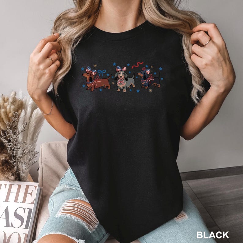 May include: Black t-shirt featuring three dachshunds with patriotic bow ties and star accents. The dogs are in shades of brown, gray, and black. The word "BLACK" is printed at the bottom right.