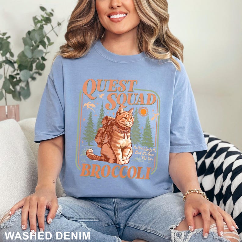 May include: A light blue t-shirt with the words "QUEST SQUAD BROCCOLI" in orange. The graphic features a cat with a backpack in a forest scene. The shirt is made of a soft material and is labeled "WASHED DENIM" at the bottom.