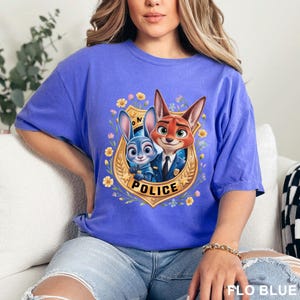 K&ouml;nnte beinhalten: Ein periwinkleblaues T-Shirt mit einer Cartoon-Grafik von Judy Hopps und Nick Wilde aus Zootopia in Polizeiuniformen. Die Grafik befindet sich in einem goldenen Abzeichen mit dem Wort "POLICE". Das Shirt hat unten den Text "FLO BLUE".