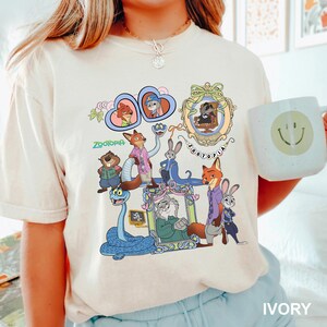 May include: Ivory-colored t-shirt featuring a colorful graphic of characters from the animated film "Zootopia." The design includes framed portraits and various animal characters. A white mug with a smiley face is held in the image.
