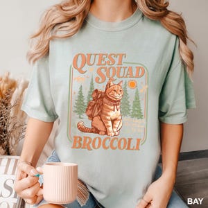 May include: A light green t-shirt with a graphic featuring a cat wearing a backpack, the words "Quest Squad Broccoli", and a forest scene. The design is in orange and green.