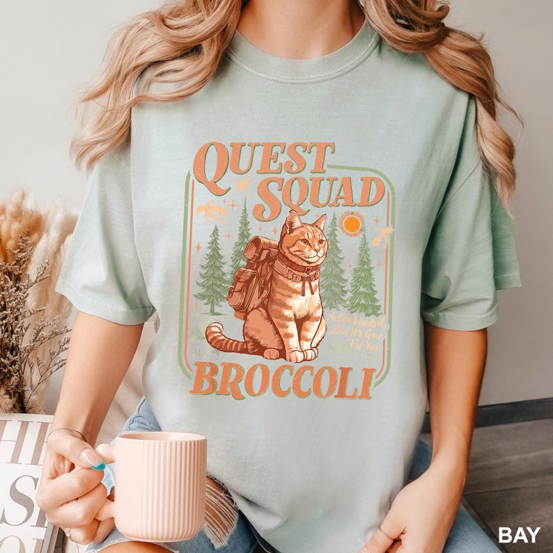 May include: A light green t-shirt with a graphic featuring a cat wearing a backpack, the words "Quest Squad Broccoli", and a forest scene. The design is in orange and green.