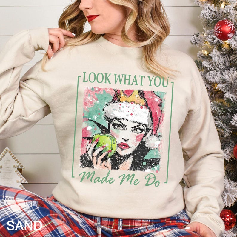 May include: Sand-colored sweatshirt featuring a graphic of a woman wearing a Santa hat and holding a green apple. The text "LOOK WHAT YOU" and "Made Me Do" are printed on the front. The sweatshirt has long sleeves and a crew neck.