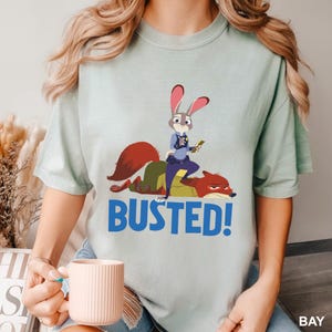 May include: A light green t-shirt featuring a cartoon image of a rabbit police officer standing on a fox, with the word "BUSTED!" in blue. The rabbit is holding a small object. The shirt is a relaxed fit.