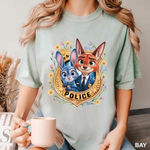K&ouml;nnte beinhalten: Hellgr&uuml;nes T-Shirt mit einer Cartoon-Illustration eines blauen Kaninchens und eines roten Fuchses in Polizeiuniformen, umgeben von einem goldenen Abzeichen mit dem Wort "POLICE" und floralen Akzenten. Das Shirt ist aus weichem Stoff.