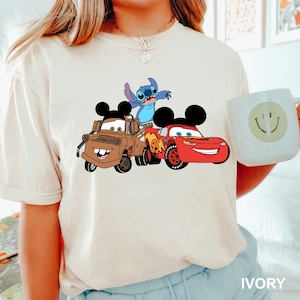 May include: A white t-shirt with a graphic print of Stitch from Lilo & Stitch, Mater from Cars, and Lightning McQueen from Cars. The characters are wearing Mickey Mouse ears.