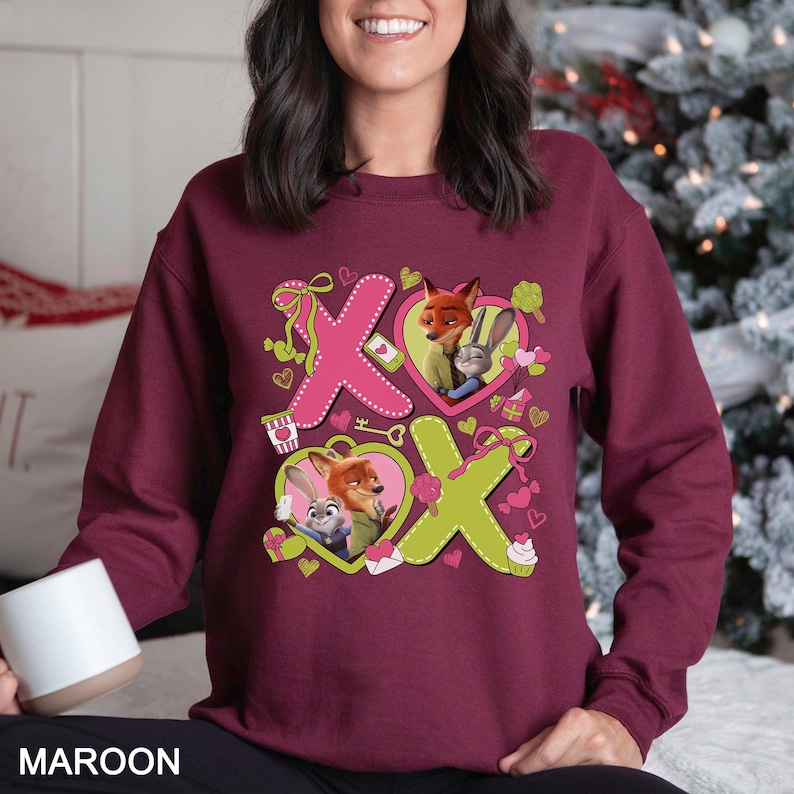 May include: Maroon sweatshirt featuring the characters Nick Wilde and Judy Hopps from Zootopia. The design includes pink and green X's with heart-shaped illustrations of the characters, along with small hearts and gift boxes.